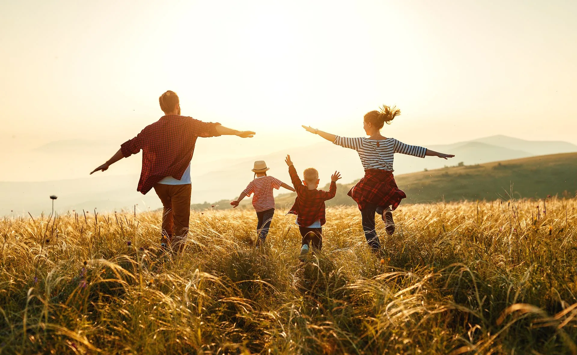 Expat family - parents and two children - running through a sunlit Swiss meadow at sunset with mountains in the distance